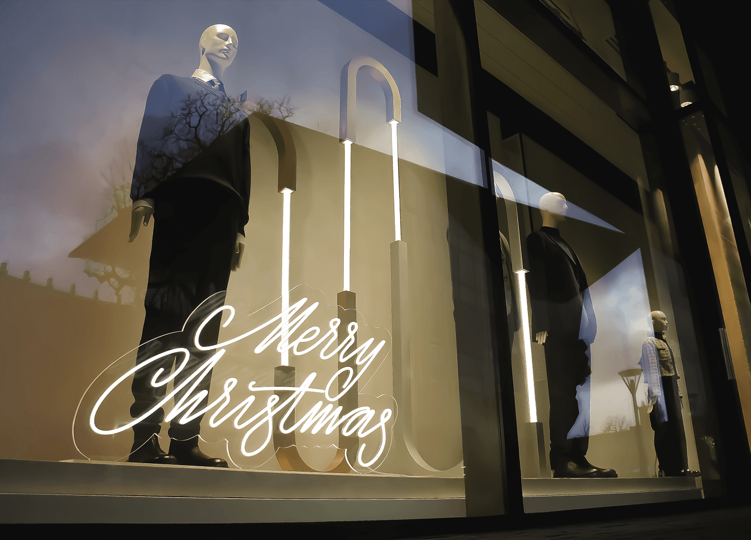 A complete Christmas window display created by PRETENDE. The exterior view shows a lit “Merry Christmas” sign on a base, vertical decorative elements and a collection displayed on mannequins.